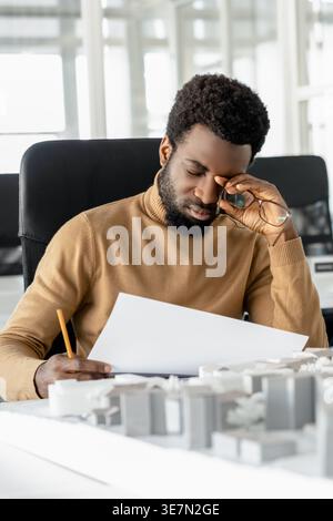 Müder afroamerikanischer Architekt, der Baupläne im Büro überprüft Stockfoto