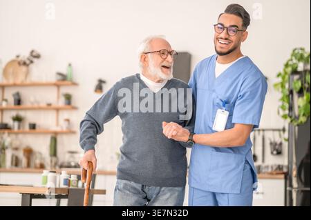 Lächelnder arabischer männlicher Betreuer in Brille, der einen älteren Mann im Stehen zu Hause hält Stockfoto