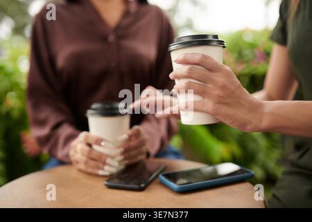 Zugeschnittenes Bild von gemischten und asiatischen Freundinnen, die Einwegkaffeetassen halten, während sie mit dem Smartphone draußen am Tisch sitzen Stockfoto