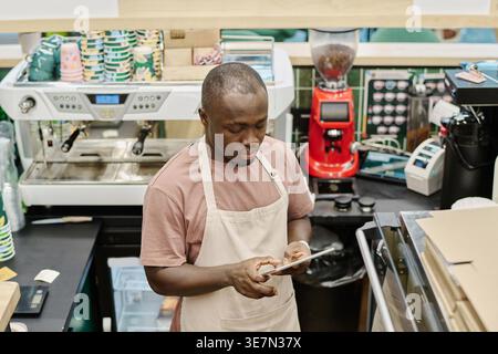 Schwarzer männlicher Ladenarbeiter mit Schürze und Tablette im Lebensmittelgeschäft Stockfoto