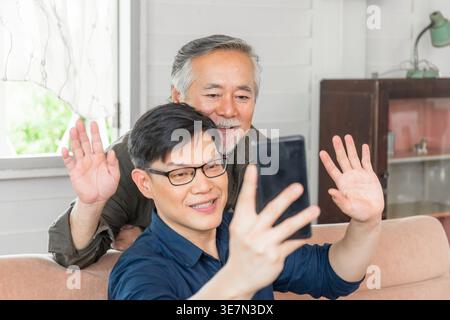 Asiatischer älterer Vater, der hinter einem Mann mittleren Alters steht und ein Smartphone benutzt, das auf der Couch sitzt, während beide im Wohnzimmer zu Hause Videoanrufe führen Stockfoto