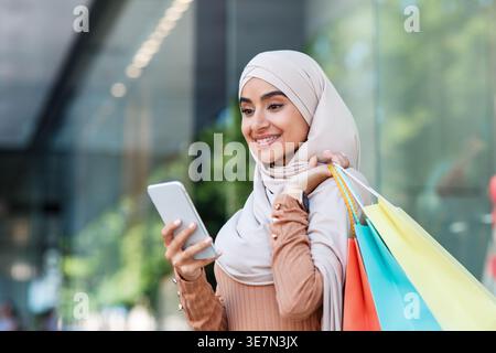 Lächelnde indische muslimische Frau, die tagsüber einen Hijab trägt und Einkaufstaschen auf der Straße in der Nähe des Einkaufszentrums trägt Stockfoto