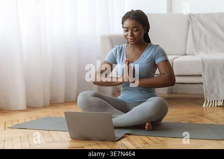 Schwarze Frau mit Online-Meditations-Yoga-Kurs, die in Namaste vor Laptop im Wohnzimmer zu Hause sitzt Stockfoto