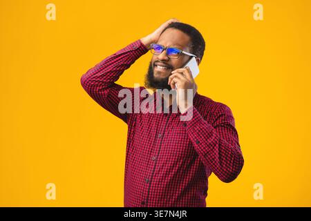 Verärgerter Schwarzer Mann mit Brille, der einen Anruf hat und wegschaut, während er vor gelbem Hintergrund im Studio steht Stockfoto