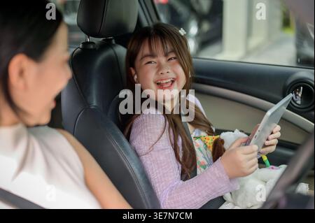 Fröhliches Thai-Mädchen, das ein Tablet hält und Spaß hat, mit ihrer Mutter zu plaudern, während sie morgens auf dem Weg zur Schule im Auto sitzt Stockfoto