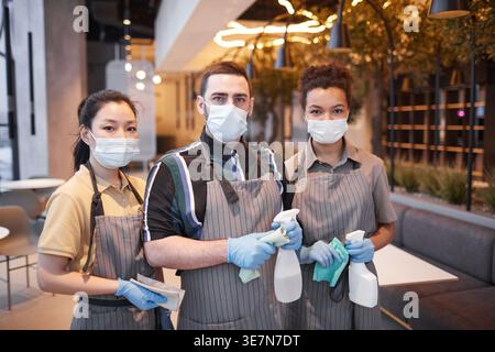Gruppe von Kellnern mit Gesichtsmasken und Schürzen, die Reinigungsmaterialien halten, während sie während der Pandemie zusammen im Café stehen Stockfoto