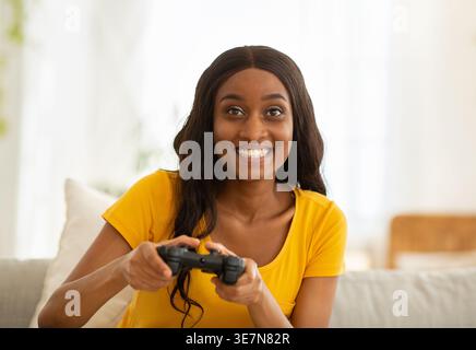 Schwarze Frau mit langen Haaren, die Videospiele mit dem Controller spielt, während sie zu Hause auf der Couch sitzt Stockfoto