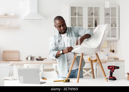 Schwarzer Mann, der Plastik auf Stuhl entfernt und Möbel auspackt, die zu Hause in der Küche stehen Stockfoto