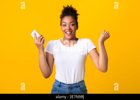 Schwarze Frau, die ein Smartphone hält, mit geballten Fäusten, die im Studio vor gelbem Hintergrund stehen Stockfoto