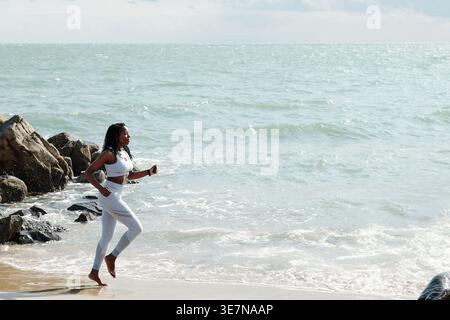 Schwarze Frau mit Dreadlocks, die ein Fitness-Outfit trägt und an sonnigen Tagen am Meer joggen Stockfoto