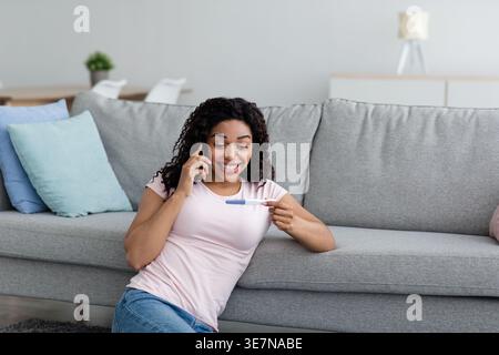 Fröhliche Schwarze Frau, die Telefongespräche führt und sich den Schwangerschaftstest ansieht, sitzt auf dem Boden vor dem Sofa im Wohnzimmer zu Hause Stockfoto