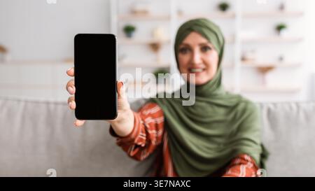 Eine muslimische Frau mittleren Alters im Hijab zeigt ein Smartphone mit leerem Bildschirm, während sie zu Hause auf dem Sofa sitzt Stockfoto