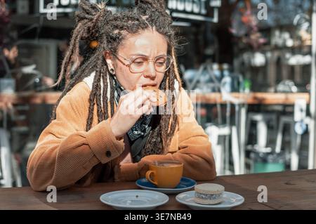 Hispanische Frau mit Dreadlocks, die eine Brille trägt und Kekse isst, die tagsüber am Tisch mit einer Kaffeetasse im Café im Freien saß Stockfoto