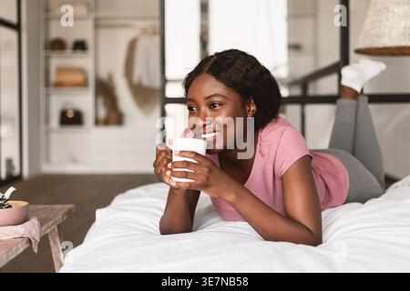 Schwarze Frau, die eine Tasse Kaffee hält und wegschaut, während sie sich im Schlafzimmer zu Hause auf dem Bett liegend Stockfoto