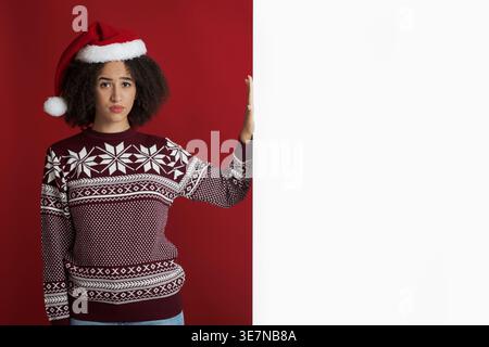 Schwarze Frau mit Weihnachtsmütze, die neben weißem Banner vor rotem Hintergrund im Studio steht Stockfoto