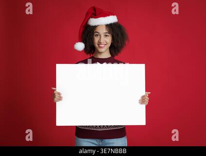 Schwarze Frau mit Weihnachtsmann-Hut mit weißem Banner, während sie vor rotem Hintergrund im Studio steht Stockfoto