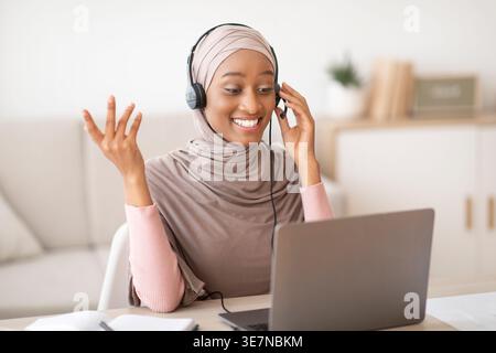 Schwarze muslimische Geschäftsfrau mit Videoanruf auf Laptop, die zu Hause am Tisch im Wohnzimmer sitzt Stockfoto