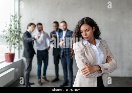 Hispanische Frau mit lockigem Haar, die über die Schulter blickt, mit überkreuzten Armen vor männlichen Kollegen, die in der Lobby im Büro sprechen Stockfoto