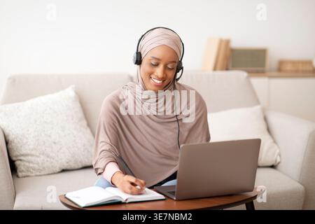 Schwarze muslimische Geschäftsfrau mit Headset, die Notizen vor dem Laptop zu Hause schreibt Stockfoto