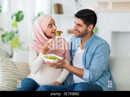 Ehemann aus dem Nahen Osten hält eine Schüssel mit Gemüsesalat und schenkt seiner schwangeren Frau aus dem Nahen Osten, während er zu Hause auf dem Sofa sitzt Stockfoto
