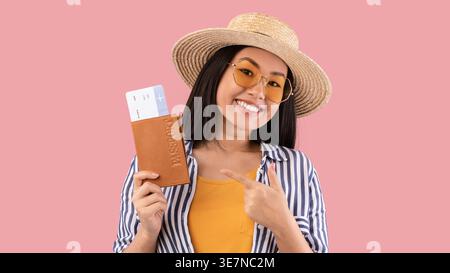Lächelnde asiatische Frau mit Strohhut und Sonnenbrille, die ... mit Flugtickets auf den Pass zeigte und auf den rosafarbenen Hintergrund stand Stockfoto