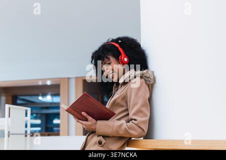 Gemischte Frau mit lockigen Haaren, die rote Kopfhörer trägt, die Buch liest, während sie sich in der Bibliothek an die Wand lehnt Stockfoto