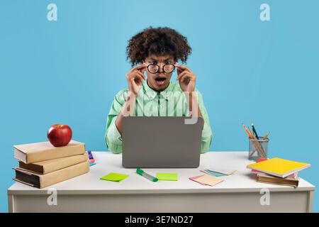 Schockierte schwarze Studentin mit lockigen Haaren und Brillen mit Laptop, der für College-Prüfungen studiert und am Tisch mit einem Stapel Bücher vor ... sitzt Stockfoto