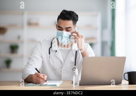 Männlicher Arzt aus dem Nahen Osten mit Schutzmaske und weißem Gewand mit Stethoskop telefoniert und Notizen macht, während er am Tisch ... sitzt Stockfoto