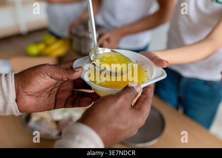 Hände des Schwarzen Mannes, der eine Schüssel Suppe hält, die von einer Gruppe Freiwilliger gegeben wurde, während er in der Mitte steht Stockfoto