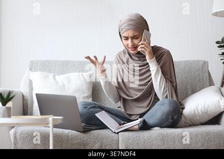 Enttäuschte arabische muslimische Frau im Hijab, die Hand im Handumdrehen beim Telefonieren mit überkreuzten Beinen auf der Couch mit Laptop und Notebook zu Hause sitzt Stockfoto