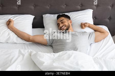 Ein arabischer Mann, der die Arme ausdehnt, während er zu Hause im Bett liegt Stockfoto