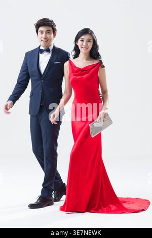 Lächelnde chinesische Frau in rotem elegantem Kleid, die Tasche hält, die mit gemischtem Rassenmann im formalen Anzug vor weißem Hintergrund im Studio geht Stockfoto