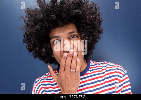 Überraschter afroamerikanischer Mann mit Hand, die den Mund bedeckt, steht an der blauen Wand Stockfoto