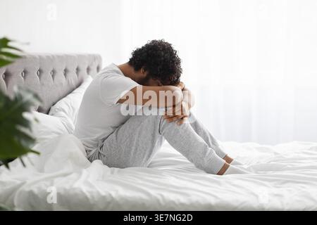 Deprimierter indischer Mann mit lockigem Haar, der Gesicht auf den Knien lehnt und auf dem Bett im Schlafzimmermattenhaus sitzt Stockfoto