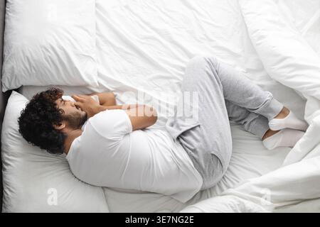 Bärtiger indischer Mann mit lockigen Haaren in weißem Hemd und Pyjama, der zu Hause im Bett liegt Stockfoto