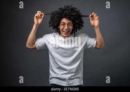 Schwarzer Mann, der eine Brille trägt und Haare zieht, während er im Studio vor dunkelgrauem Hintergrund steht Stockfoto