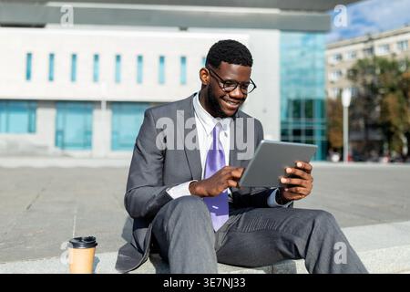 Schwarzer Geschäftsmann in Brille mit Tablet, während er an sonnigen Tagen auf der Treppe neben der Kaffeetasse zum Mitnehmen vor dem Bürogebäude sitzt Stockfoto