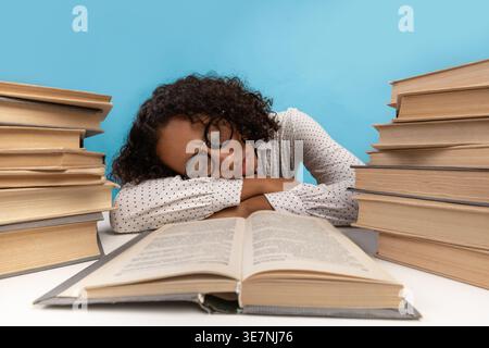 Müde schwarze Teenagerin, die eine Brille mit geschlossenen Augen trägt, sitzt und lehnt sich auf einem Tisch mit Büchern vor blauem Hintergrund im Studio Stockfoto