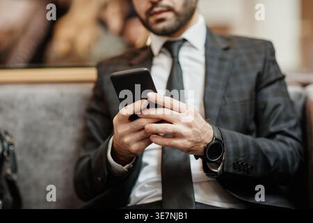 Zugeschnittenes Bild eines Geschäftsmannes aus dem Nahen Osten, der ein Smartphone verwendet, das in der Hotellobby auf einem Sessel sitzt Stockfoto