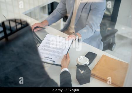 Zugeschnittenes Bild eines asiatischen Geschäftsmannes, der am Laptop arbeitet und Dokumente zu einer Kollegin bringt, während er am Tisch im Büro sitzt Stockfoto