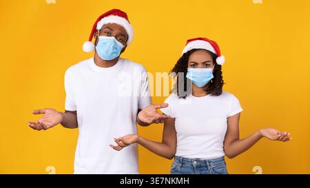 Schwarzes Paar mit Sant Hut und Schutzmaske, stehend mit offenen Armen vor gelbem Hintergrund im Studio Stockfoto