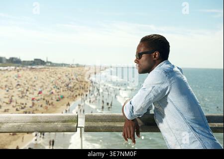 Ernster Schwarzer Mann, der wegschaut und eine Sonnenbrille trägt, die sich auf einem Geländer stützt und auf dem Balkon am Strand steht Stockfoto
