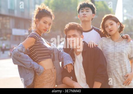 Ernsthafter chinesischer Mann, der mit Freunden auf der Straße sitzt, die an sonnigen Tagen um ihn herum stehen Stockfoto