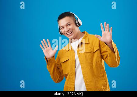 Lächelnder asiatischer Mann mit Kopfhörern, der Musik mit erhobenen Händen im Studio vor blauem Hintergrund lauscht Stockfoto