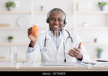 Schwarzer männlicher Arzt, der ein Headset trägt, das eine Flasche Pillen zeigt, während er in der Klinik mit Tablet und Brille am Schreibtisch sitzt Stockfoto