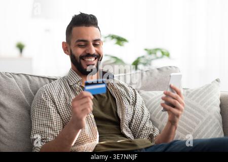 Fröhlicher arabischer Mann mit Bart, der Kreditkarte und Smartphone hält, während er zu Hause auf dem Sofa sitzt Stockfoto