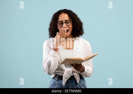 Müde schwarze Teenager mit Brille und Gähnen, während sie im Studio vor blauem Hintergrund stehen Stockfoto