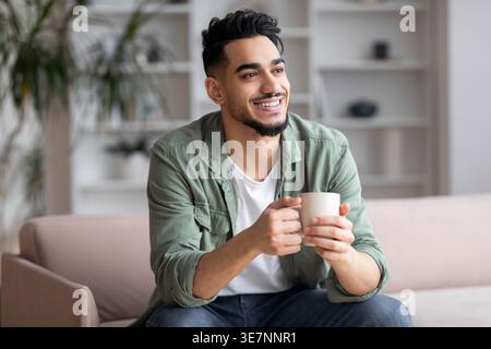 Lächelnder arabischer Mann, der wegblickt und eine Tasse Kaffee hält, der auf dem Sofa sitzt Stockfoto