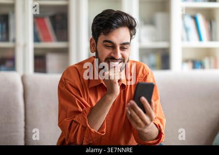 Mann aus dem Nahen Osten mit geschlossener Faust in der Nähe des Kinns und Blick auf das Smartphone, das zu Hause auf der Couch sitzt Stockfoto