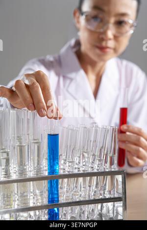 Ernsthafte chinesische Wissenschaftlerin in Schutzbrille, die Reagenzgläser hält, während sie am Tisch vor grauer Wand steht Stockfoto